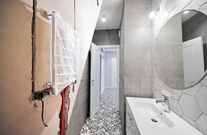 bagno prima e dopo la ristrutturazione. Nel prima si vede un muro spaccato e nel dopo una bagno nuovo con piastrelle bianche e grigie un lavandino, lo specchio grande rotondo
